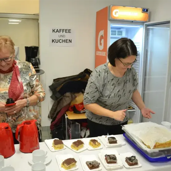 Zwei Frauen bereiten Essen auf einem Tisch in einem Küchenbereich zu, mit einem offenen Kühlschrank und einer Kaffeemaschine im Hintergrund. Ein Schild an der Wand steht 'Kaffee und Kuchen'.