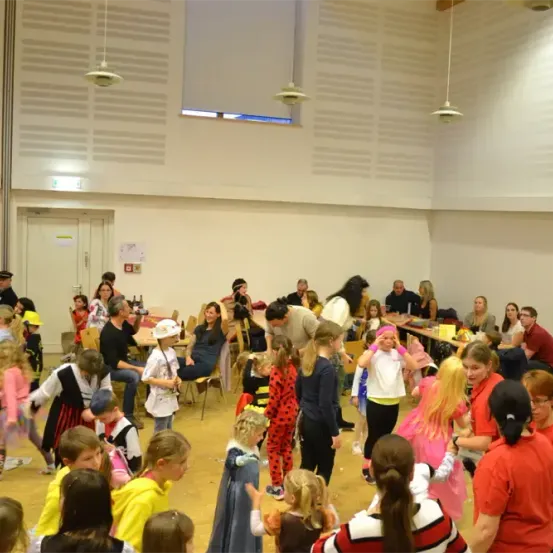 Eine Gruppe von Kindern in Kostümen tanzt in einem Saal. Mehrere Erwachsene sitzen an Tischen und einige stehen. Der Raum hat weiße Wände, hängende Lampen und ein Glasfenster.
