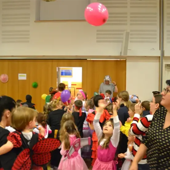 Eine Gruppe von Kindern in Kostümen ist drinnen. Ein rosa Ball schwebt in der Luft. Eine Frau in einem gepunkteten Kleid schaut zu.