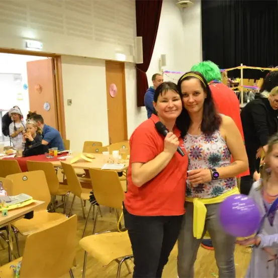 Zwei Frauen und ein Mädchen stehen in einem Raum mit Luftballons und lächeln. Die Frauen halten ein Mikrofon und einen lila Ballon. Dahinter stehen eine Person mit grünen Haaren und andere. Tische mit Stühlen und Dekorationen sind aufgestellt.