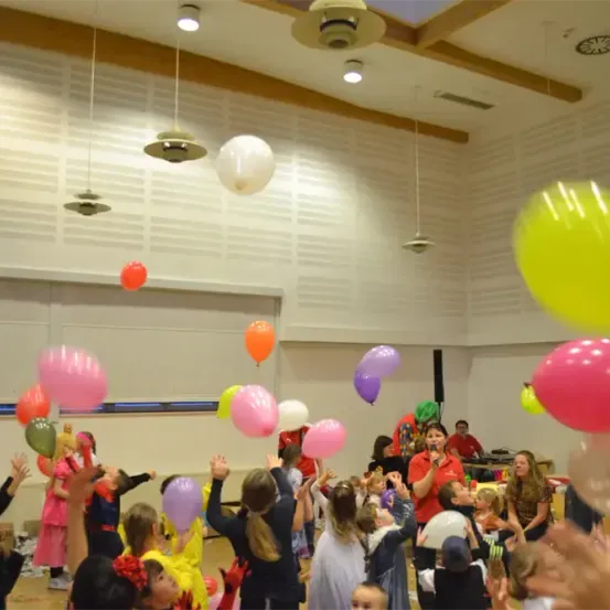 In einem Raum voller Luftballons spielen Kinder und Erwachsene und haben Spaß. Einige halten Luftballons, während andere in der Luft schweben. Eine Frau singt und hält ein Mikrofon.