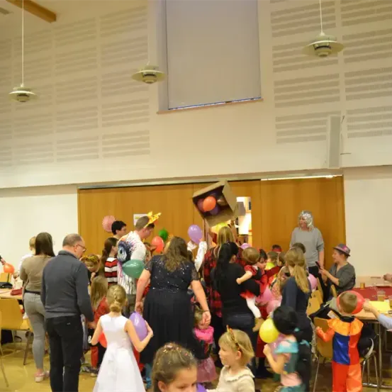 Eine große Gruppe von Menschen, einschließlich Kindern und Erwachsenen, versammelt sich drinnen. Viele sind in Kostümen gekleidet und einige halten Luftballons. Tische sind mit Stühlen aufgestellt und Menschen stehen und unterhalten sich.