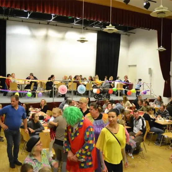 Ein großer Innenraum mit Bühne zeigt Menschen, die an Tischen essen, einer als Clown verkleidet, mit bunten Luftballons und Girlanden. Der Raum hat Holzböden und eine hohe Decke mit Beleuchtung.