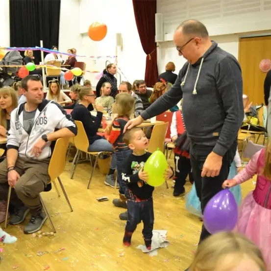 Eine Gruppe von Menschen feiert drinnen einen Geburtstag. Ein Mann hält einen Luftballon für einen kleinen Jungen. Andere Kinder stehen herum. Der Raum ist mit Luftballons und Bändern dekoriert.