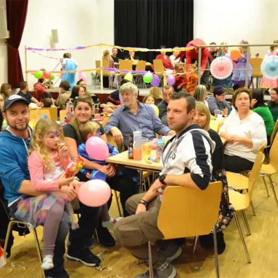 Eine große Gruppe von Menschen versammelt sich in einem festlichen Ambiente, geschmückt mit Luftballons und Girlanden. Erwachsene und Kinder sitzen an Tischen, manche halten Luftballons. Die Stimmung ist lebhaft, mit Menschen, die sich unterhalten und die Veranstaltung genießen.