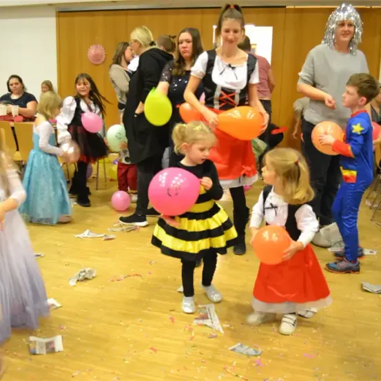 Eine Gruppe von Kindern und Erwachsenen feiert mit Luftballons in einem Raum, einige in Kostümen. Ein junges Mädchen in einer Bienenkostüm hält einen rosa Luftballon.