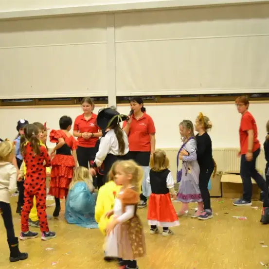 Eine Gruppe von Kindern in Kostümen versammelt sich in einem Raum mit Erwachsenen. Einige Kinder stehen, andere sitzen oder gehen. Ein Kind hält ein Mikrofon.