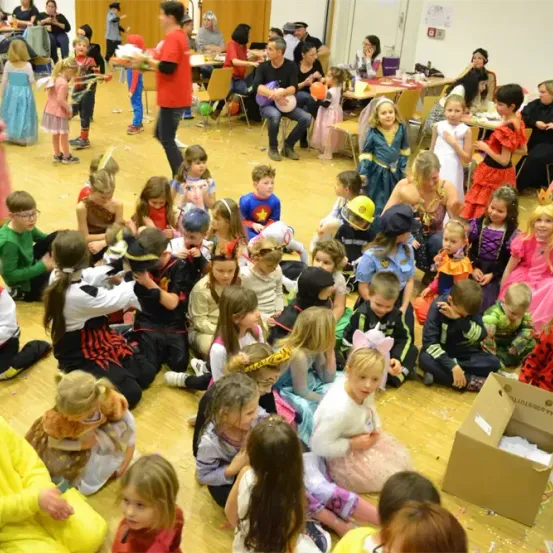 Eine Gruppe von Kindern in verschiedenen Kostümen sitzt und steht auf einem Holzboden. Einige essen, während andere etwas betrachten. Eine Person in einem roten Shirt steht in der Nähe eines Stuhls. In der Ferne sitzen Leute auf Stühlen und Tischen, mit Luftballons darauf. Es gibt auch Poster an der Wand.