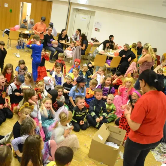 Eine Gruppe von Kindern in Kostümen sitzt auf dem Boden in einem Raum. Eine Frau steht vor einer Pappschachtel und scheint sie anzusprechen. Tische und Stühle sind im Hintergrund aufgestellt.