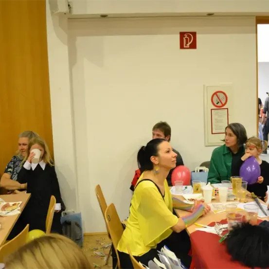 Eine Gruppe von Menschen sitzt um einen Tisch mit Essen und Getränken. Eine Frau in einem gelben Shirt schaut etwas an. Hinter ihr befindet sich ein Ballon und ein Schild an der Wand.