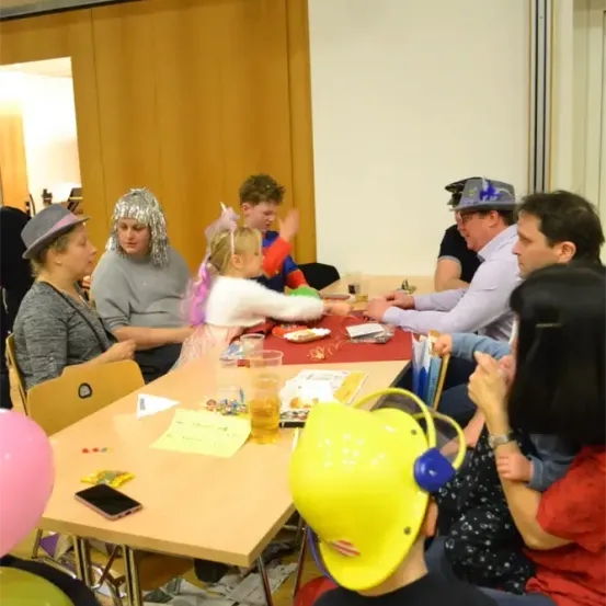 Eine Gruppe von Menschen, darunter Kinder, sitzt um einen Tisch herum mit Luftballons, Bechern und Papieren. Einige tragen Hüte, während andere Perücken haben. Sie scheinen sich in einem Raum zu befinden, möglicherweise während einer Party.