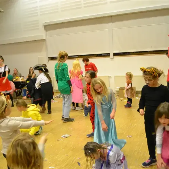 Eine Gruppe von Kindern in verschiedenen Kostümen tanzt in einem Raum, einige halten sich an den Händen. Eine Person in einem roten Kostüm ist in der Nähe. Eine weiße Wand und eine weiße Decke sind sichtbar.