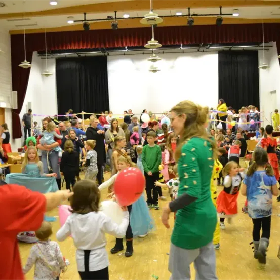 Eine Gruppe von Menschen, hauptsächlich Kinder, befindet sich in einem Saal und nimmt an einer Kostümparty teil. Einige halten Luftballons. Eine Frau in Grün hält einen roten Ballon.
