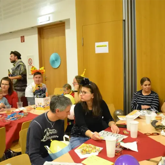 Eine Gruppe von Menschen sitzt an Tischen in einem Raum, einige tragen Hüte, Brillen und lächeln. Sie feiern eine Party mit Luftballons, Papiertellern und Bechern. Ein Mann hält eine Flasche. Hinter ihnen befinden sich zwei Türen und eine Wand mit einem Papierschild.
