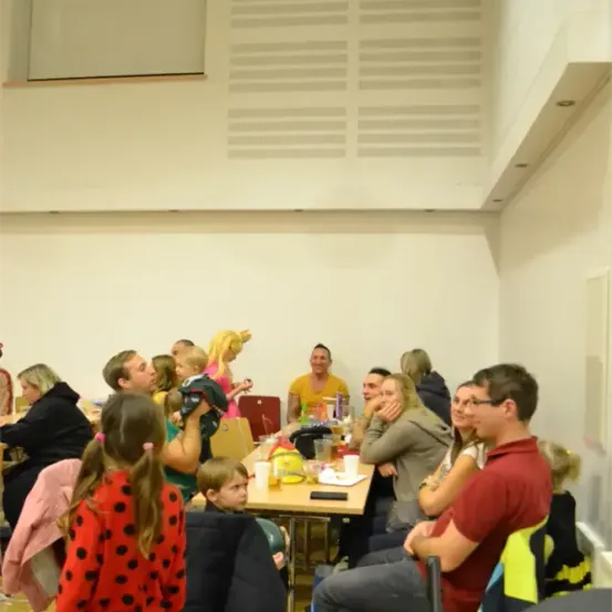 Eine Gruppe von Menschen, darunter Erwachsene und Kinder, sitzen an Tischen in einem Raum. Einige essen, während andere reden. Ein Mädchen in einem roten Tupfenkleid steht vor einem Tisch.