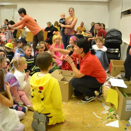 Eine Frau mit einem Mikrofon spricht eine Gruppe von Kindern an, einige in Kostümen, mit Kisten und Konfetti auf dem Boden.