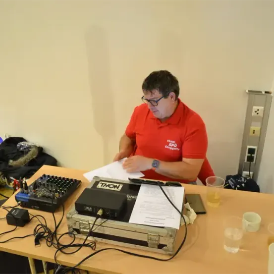 Ein Mann in einem roten Shirt sitzt an einem Schreibtisch, umgeben von Audiogeräten, und scheint konzentriert auf Papierkram zu sein.