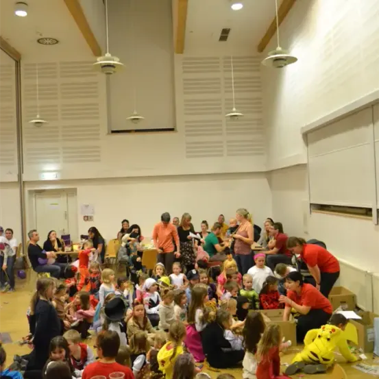 Eine Gruppe von Kindern in Kostümen sitzt auf dem Boden, einige stehen, in einem Raum mit Erwachsenen. Tische mit Essen und Getränken sind vorhanden. Einige Erwachsene stehen in der Nähe der Wand, möglicherweise organisieren sie die Veranstaltung.