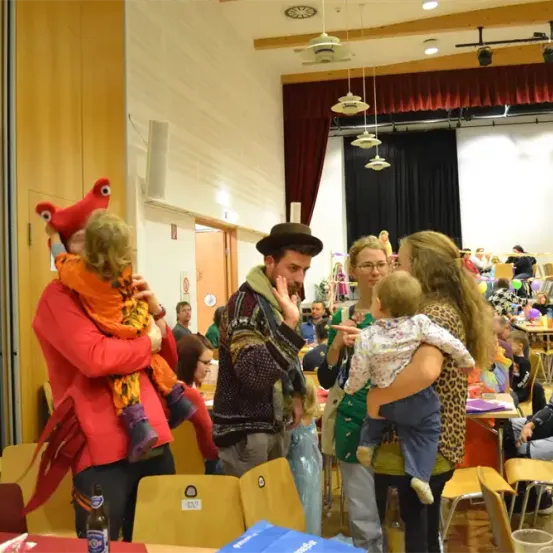 Eine Familienveranstaltung in Innenräumen mit einem Mann in einem Hut, der ein Kind hält, und einer Frau, die ein Baby hält. Andere Familien sitzen an Tischen. Die Umgebung ist ein Raum mit Holzwänden und Stühlen.