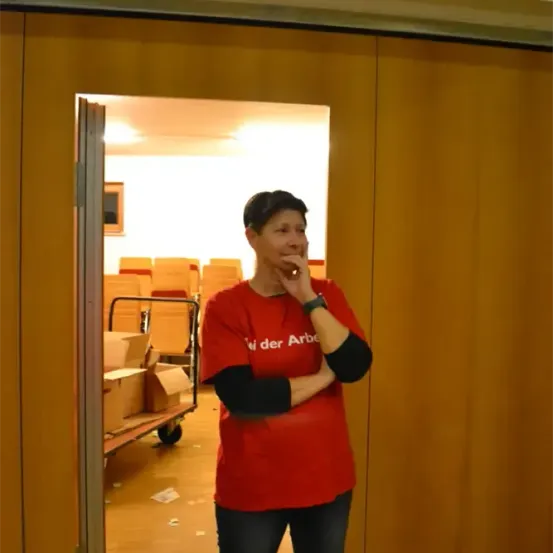 Eine Frau in einem roten T-Shirt mit den Worten 'Wir der Arbeit' steht in einer offenen Tür, lächelt und telefoniert. Hinter ihr befindet sich ein Raum mit Stühlen und Kisten.