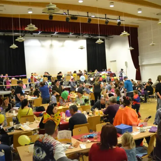 Ein großer Saal voller Menschen bei einem Festival. Viele sitzen an Tischen, einige stehen, alle tragen bunte Kostüme und Hüte. Überall sind Luftballons und Dekorationen. Die Bühne ist mit Vorhängen und Lichtern ausgestattet.