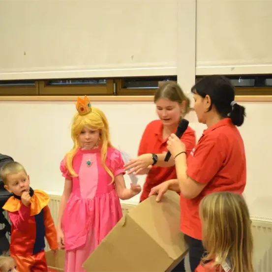 Eine Frau hält ein Mikrofon und spricht eine Mädchen in einem rosa Kleid an. Eine andere Mädchen hält eine Box. Kinder beobachten, einige in Kostümen. Ein Kinderwagen steht in der Nähe.