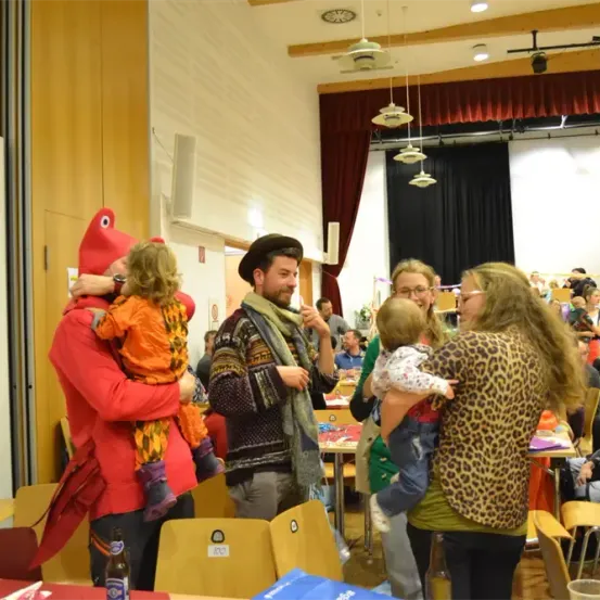 In einem großen Raum sind mehrere Erwachsene und Kinder versammelt, wahrscheinlich für eine Veranstaltung. Ein Erwachsener hält ein Kind in einem roten Kostüm. Ein anderer Erwachsener hält ein Baby und steht neben einer Frau in einem Leopardenprint-Shirt. Der Raum hat Tische, Stühle und eine Bühne mit Vorhängen.