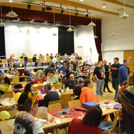 Ein großer Saal mit Tischen und Stühlen, der ein festliches Treffen beherbergt. Menschen sitzen an Tischen, einige stehen. Dekorationen umfassen Luftballons und Banner. Scheinwerfer hängen von der Decke ab.