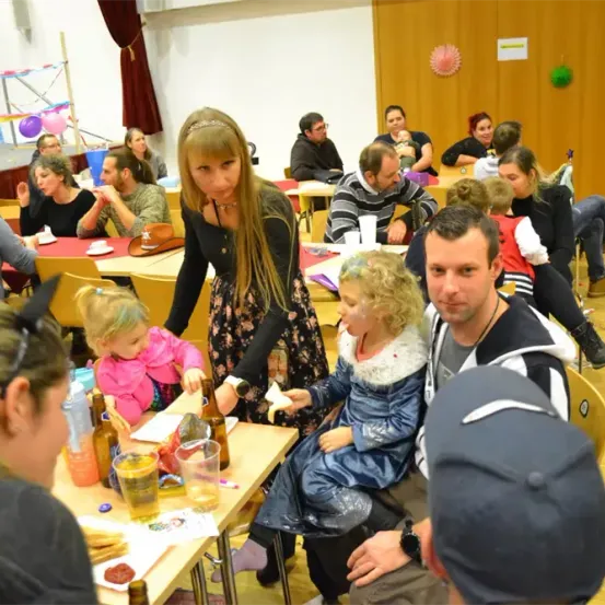 Eine festliche Zusammenkunft im Innenbereich, bei der Erwachsene und Kinder an Tischen sitzen. Eine Frau in einem Blumenkleid serviert Essen, während ein Mädchen in einem blauen Kleid ein Stück Brot hält.