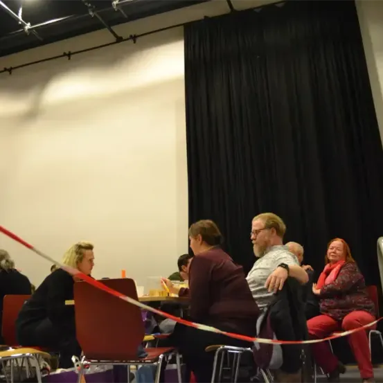 Mehrere Personen sitzen um Tische herum, mit einer roten Bandbarriere davor. Der Bereich hat eine Bühne mit schwarzem Vorhang.