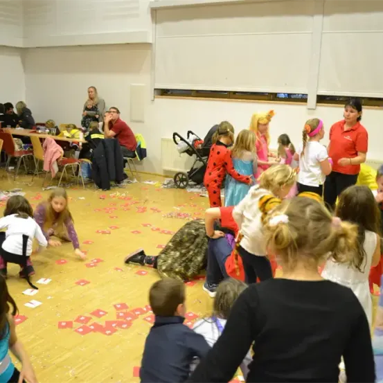 Kinder spielen auf dem Boden mit roten Karten, und einige tragen Kostüme. Einige Erwachsene stehen und beobachten. Tische und Stühle sind im Raum aufgebaut.