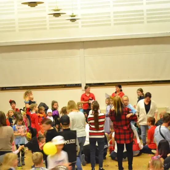Eine Gruppe von Menschen, darunter Kinder, befindet sich in einem Raum. Einige stehen, andere sitzen auf dem Boden. Es gibt Erwachsene in roten Shirts und einen Mann in einem schwarzen Shirt mit dem Wort 'POLIZEI' darauf. Es gibt Luftballons und einige Spielzeuge auf dem Boden.