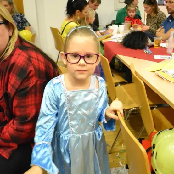 Ein Mädchen in einem blauen Kleid mit Brille steht auf einer Party. Erwachsene und Kinder sitzen an Tischen. Der Raum hat eine festliche Atmosphäre mit Luftballons und Dekorationen.