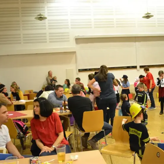 Ein großer Raum mit weißen Wänden und Tischen. Menschen sitzen in Stühlen und einige stehen. Ein Kind trägt eine Feuerwehruniform und einen gelben Helm. Flaschen und Gläser stehen auf den Tischen.