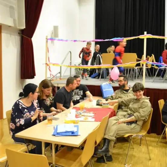Mehrere Personen sitzen an einem Tisch in einem Raum mit Bühne, Luftballons und einer Balustrade. Ein Kind in einem roten Kostüm steht auf der Bühne.