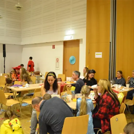 Eine Gruppe von Menschen versammelt sich in einem Raum, einige sitzen an Tischen mit Essen und Getränken, andere stehen. Es gibt Luftballons und Dekorationen. Eine Frau in einem blauen Kleid schaut sich etwas auf dem Tisch an.