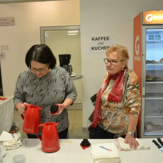 Zwei Frauen stehen an einem Tisch mit roten Kaffeekannen. Sie bereiten wahrscheinlich Kaffee für Kunden zu. Der Tisch ist mit Servietten, Tassen und Messern gedeckt. Ein Kühlschrank ist im Hintergrund zu sehen, und ein Schild mit der Aufschrift 'KAFFEE UND KUCHEN' ist angebracht.