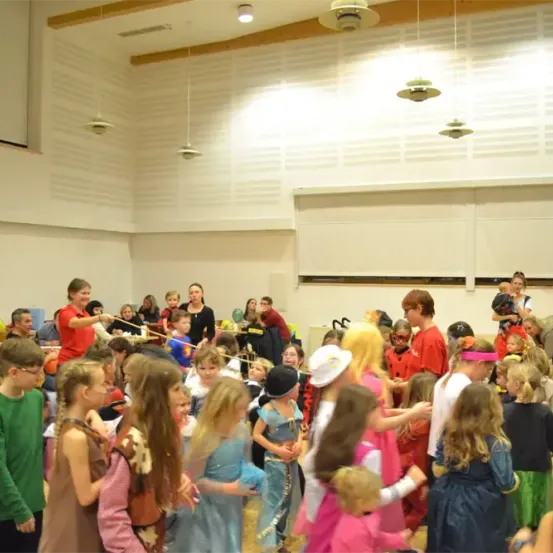 Eine Gruppe von Kindern in Kostümen und Masken tanzt in einem Raum mit weißen Wänden und Holzbalken. Einige tragen Brillen, andere haben Accessoires. Ein Mann in einem roten Hemd hält einen Stock, und mehrere Personen sind im Hintergrund.