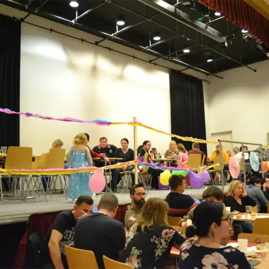 Eine festliche Veranstaltung in einem Bankettsaal mit Menschen an Tischen. Ein Bühnenbereich ist mit Luftballons und Girlanden dekoriert. Eine Frau in einem blauen Kleid steht auf der Bühne.