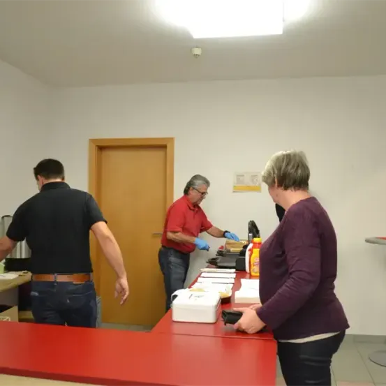 Drei Personen befinden sich in einer Küche; ein Mann in Schwarz steht in der Nähe eines Tresens, ein Mann in Rot mit Handschuhen bereitet Lebensmittel am Tresen zu, und eine Frau in Lila steht am Tresen.