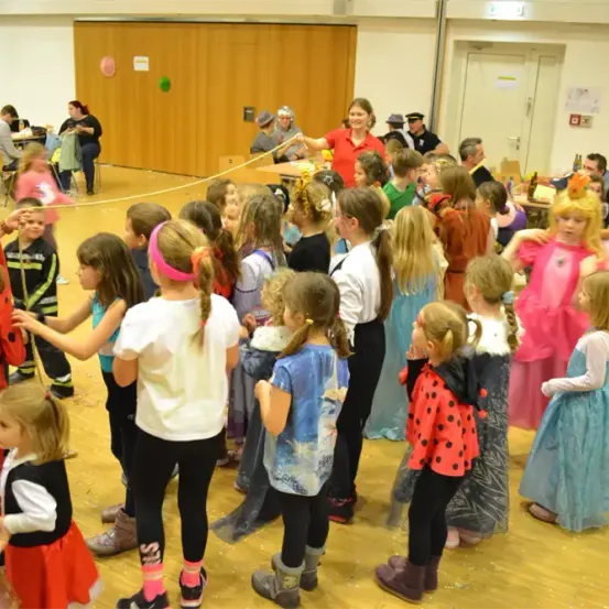 Eine Gruppe von Kindern in Kostümen tanzt in einem Saal in einem Kreis. Ein Erwachsener in Rot hält eine lange Schnur. Der Raum hat Holzböden und Wände.