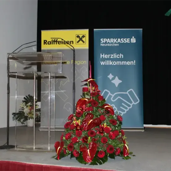 Ein Rednerpult mit Mikrofonen steht auf einer Bühne, flankiert von einer roten Blumenarrangement und Bannern für Raiffeisen und Sparkasse Neunkirchen, das 'Herzlich willkommen!' vor schwarzem Hintergrund liest.