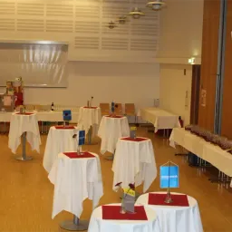 Ein Saal mit Holzböden und weißen Wänden. Mit weißen Tüchern bedeckte Tische sind aufgestellt, jeder mit einem roten Tuch, einer Blume und einem Glas. Ein blauer Banner ist im Vordergrund sichtbar.