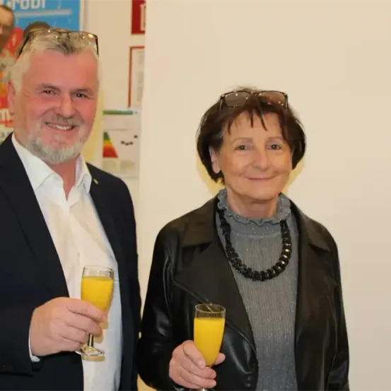 Ein älterer Mann und eine ältere Frau stehen nebeneinander und halten jeweils ein Glas Orangensaft. Sie lächeln und scheinen für ein Foto zu posieren. Hinter ihnen befindet sich eine weiße Wand mit einem Plakat.