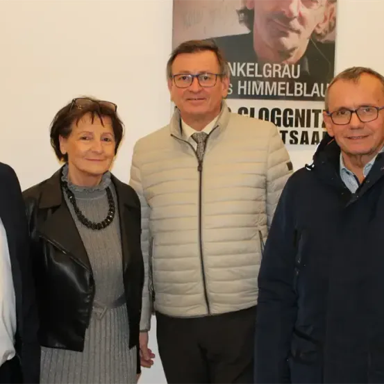 Vier Menschen stehen eng beieinander vor einer weißen Wand. Sie alle lächeln. Hinter ihnen ist ein Poster mit einem Foto eines lächelnden Mannes.