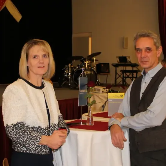 Ein Mann und eine Frau stehen neben einem Tisch mit einem Schild davor, möglicherweise bei einer Veranstaltung. Dahinter ist eine Bühne mit Musikinstrumenten zu sehen.