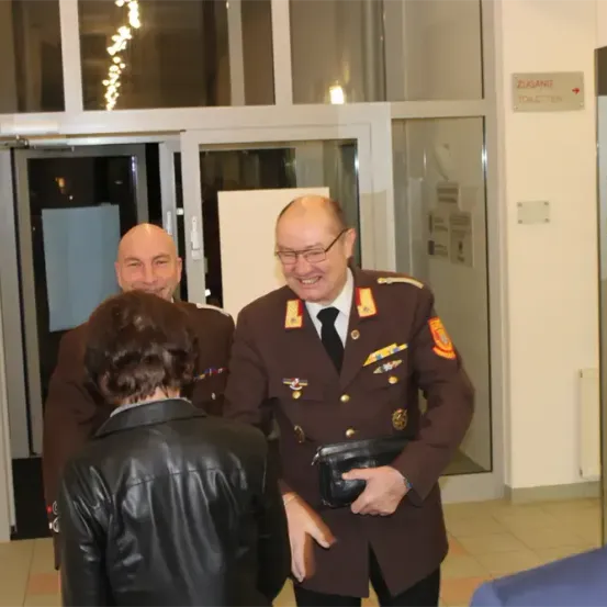 Eine Gruppe von Menschen, darunter ein Mann in Uniform, steht in einem Flur. Er hält eine schwarze Brieftasche und lächelt. Hinter ihnen befindet sich eine Glastür mit einem Schild, das 'Zugang' besagt.