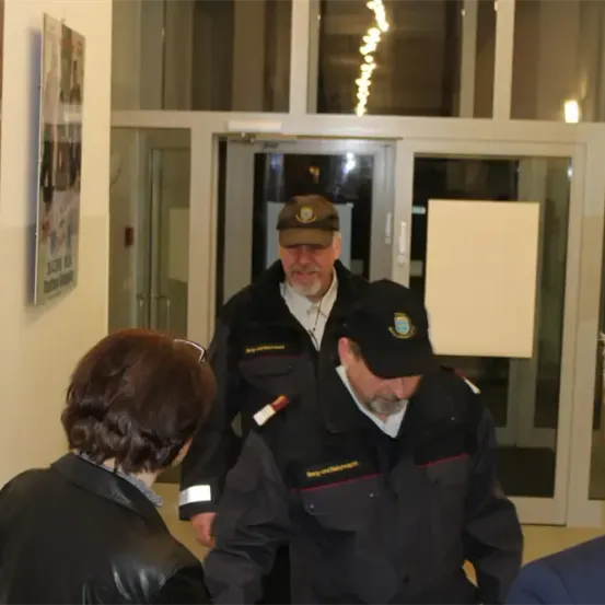 Eine Gruppe von Menschen steht in der Nähe einer Tür. Zwei Männer in schwarzen Uniformen und Mützen stehen und reden. Einer von ihnen hält ein Mikrofon. Eine Frau in einer schwarzen Jacke steht in ihrer Nähe. Es gibt Poster an der Wand.