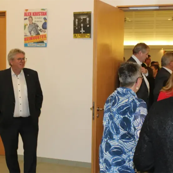 Eine Gruppe formell gekleideter Männer und Frauen versammelt sich in einem Flur und blickt auf ein Poster mit dem Namen Alex Kristan.