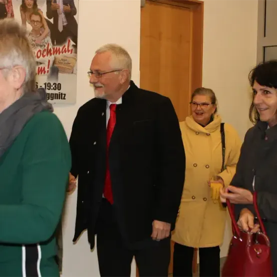 Vier Personen stehen in einem Innenraum, wahrscheinlich einem Flur. Sie scheinen ein Gespräch zu führen. Der Mann in der Mitte trägt eine rote Krawatte und eine Brille. Die Frau auf der rechten Seite hält eine rote Handtasche. Hinter ihnen befindet sich ein Plakat mit Text.